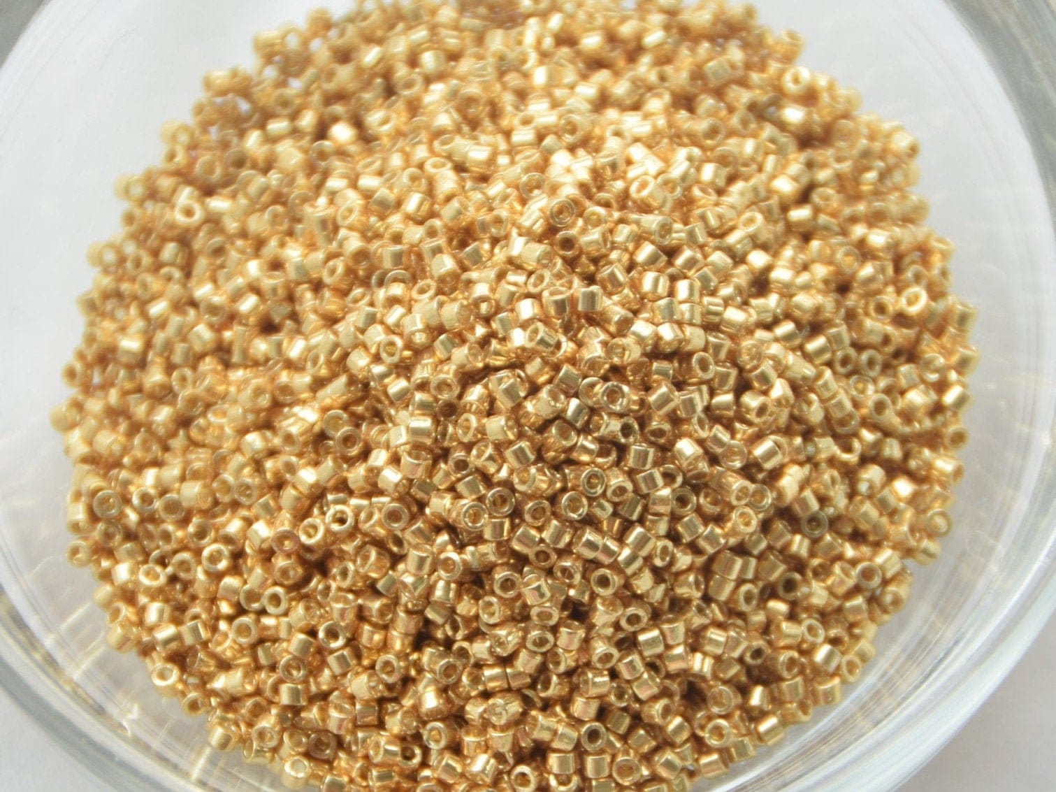 Metallic gold seed beads, 1.6mm in size and cylindrical in shape. The image is a close-up of a round bowl of full of seed beads on a white background.
Miyuki Delica beads, gold colored.