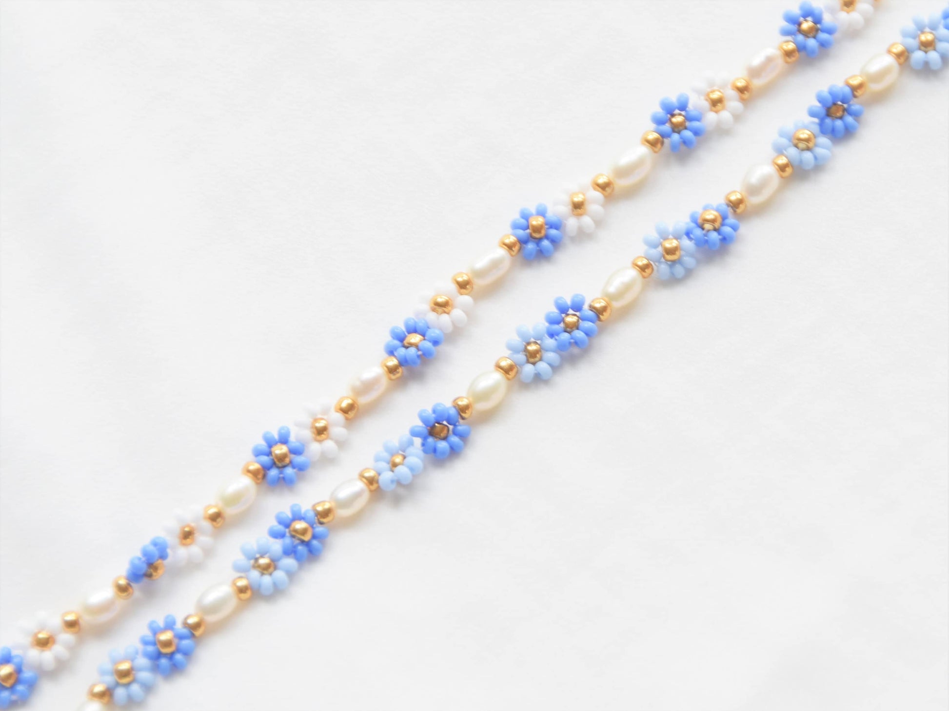 Two bracelets, one with blue and white flowers and the other one with two shades of blue flowers, lying straight, parallel to each other, diagonally across the image. Both bracelets have freshwater pearls, alternating with the flowers.