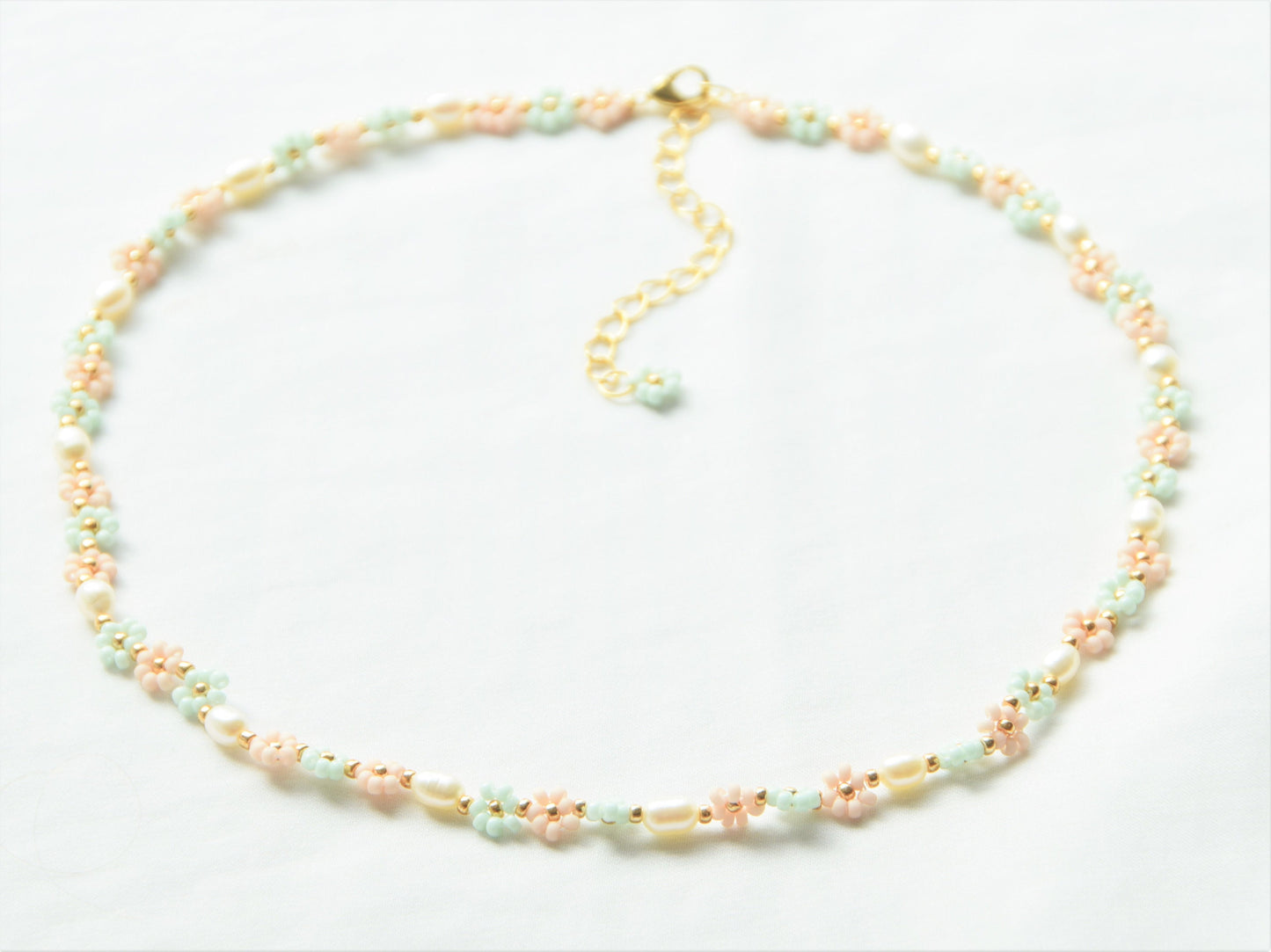 A necklace lying flat, making a circle, on a white background. The necklace has light green and light pink flowers, alternating each other in sets of three. Between each set of three flowers, is an oval shaped freshwater pearl.