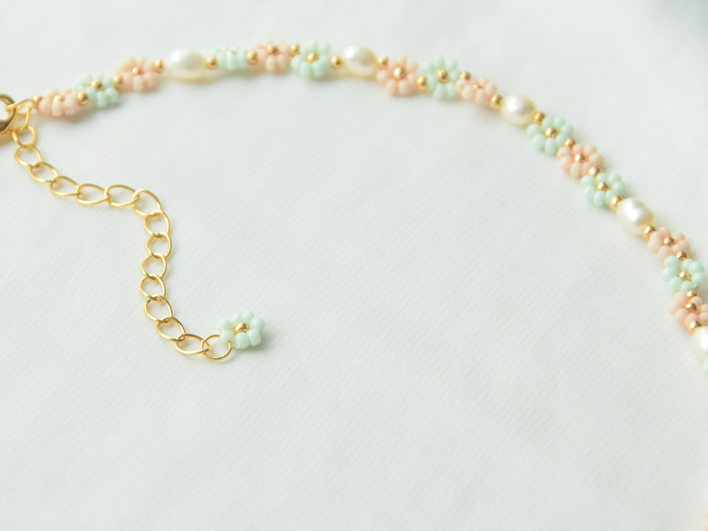 Close-up image of a necklace lying flat on a white background. The necklace has light green and light pink flowers, alternating each other in sets of three. Between each set of three flowers, is an oval shaped freshwater pearl.