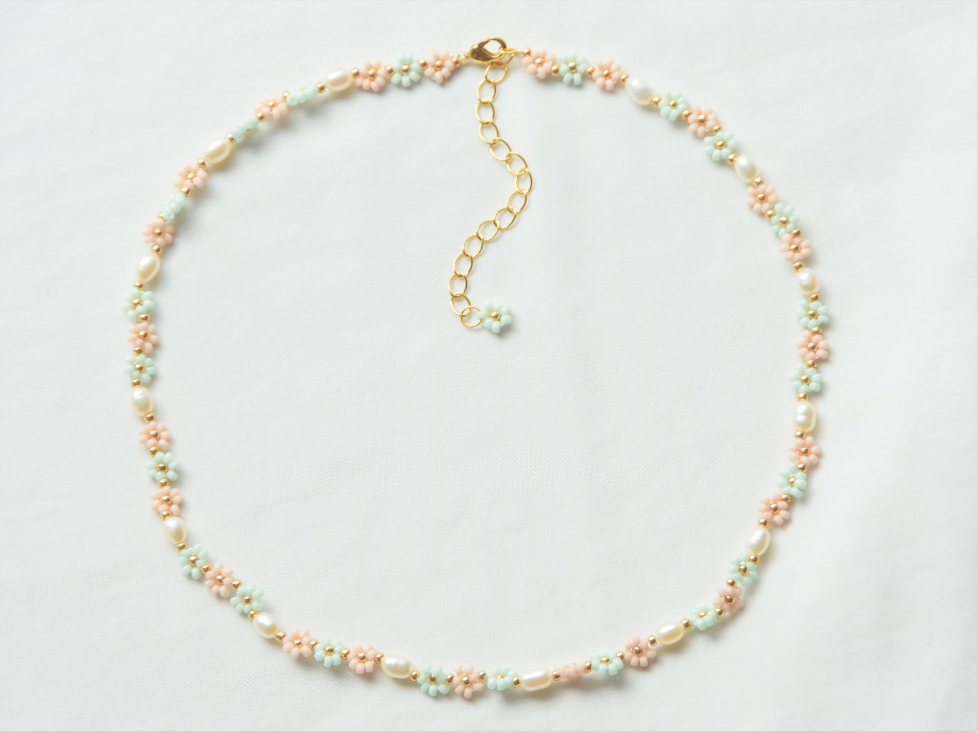 A necklace lying flat, making a circle, on a white background. The necklace has light green and light pink flowers, alternating each other in sets of three. Between each set of three flowers, is an oval shaped freshwater pearl.