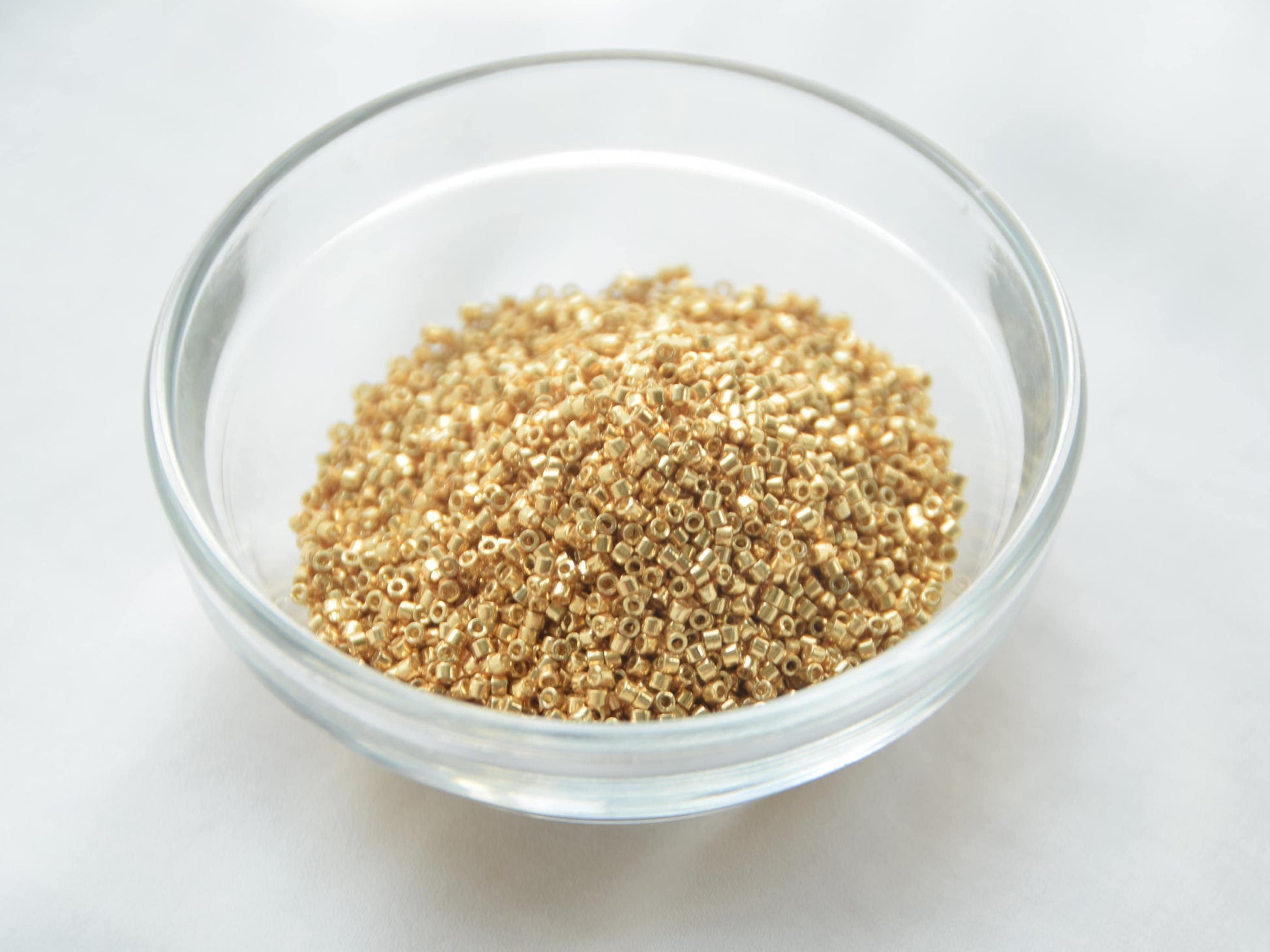 A round transparent bowl half filled with metallic gold beads, 1.6mm in size and cylindrical in shape. The bowl lies on a white background.