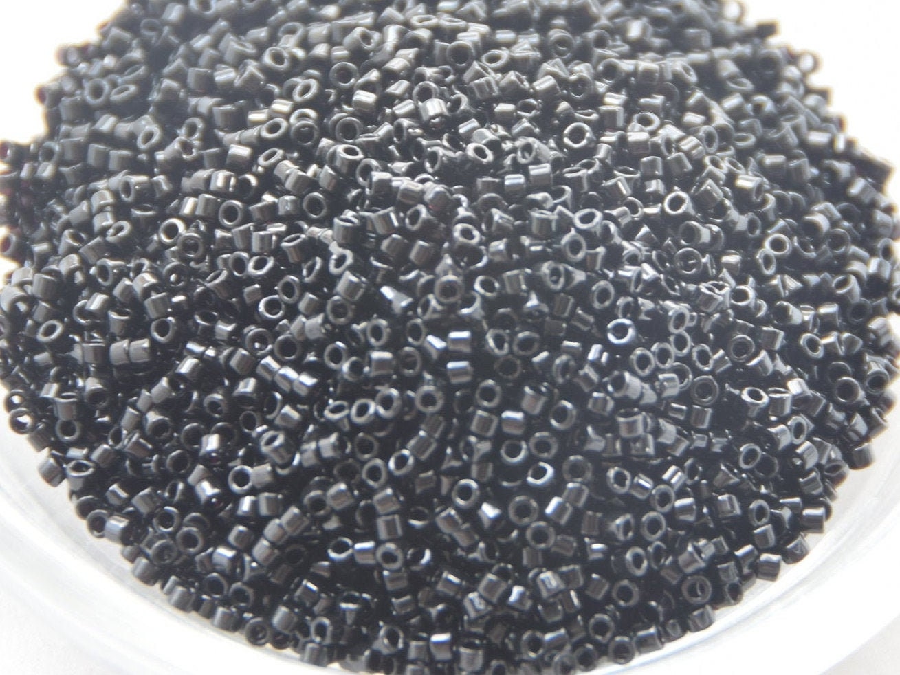 Opaque black seed beads, 1.6mm in size and cylindrical in shape. The image is a close-up of a round bowl of full of seed beads on a white background. 
Miyuki delica beads opaque black.