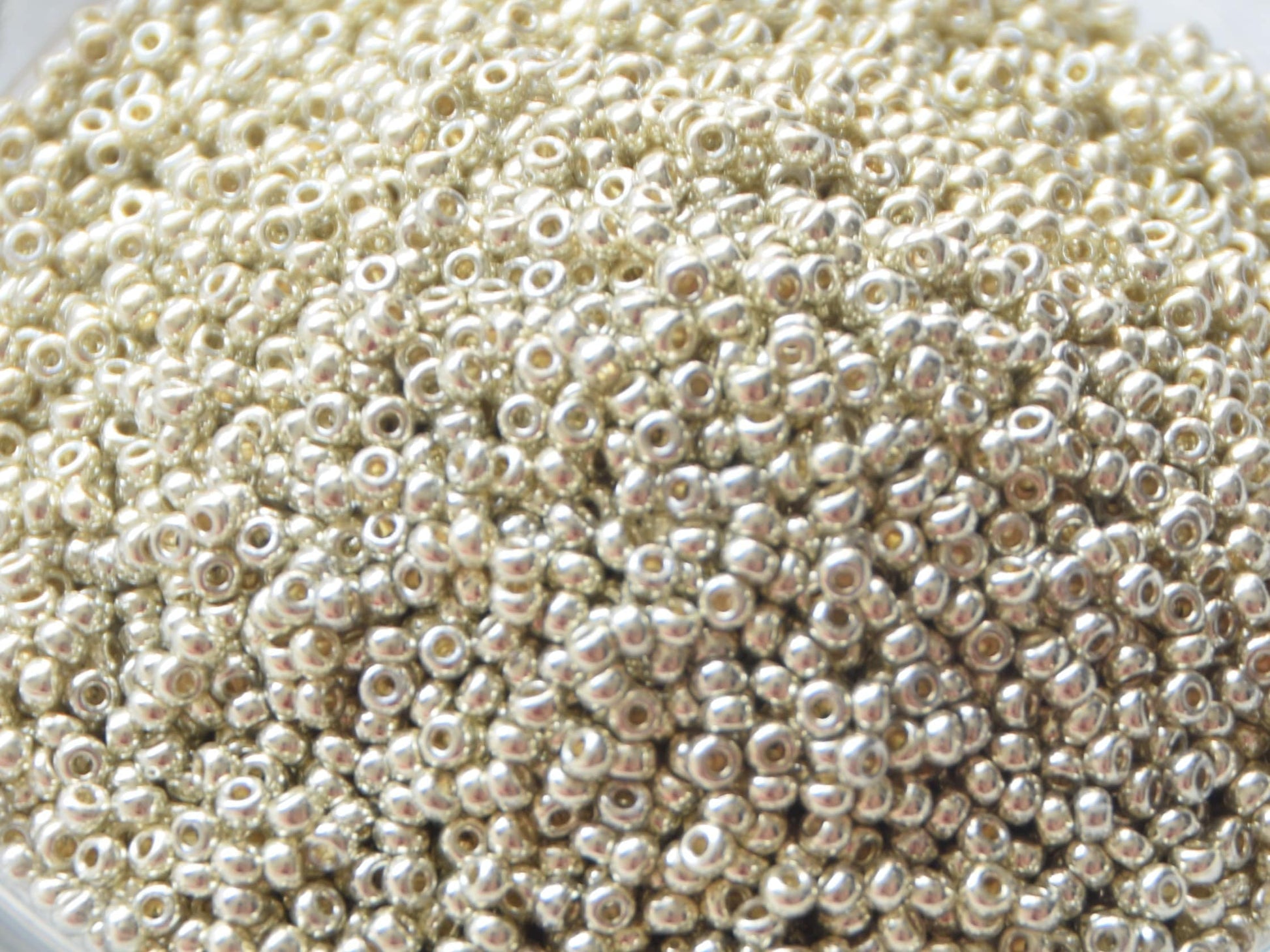 Metallic silver seed beads, 2mm in size and round in shape. The image is a close-up of a bowl of full of seed beads so that the beads fill the entire image