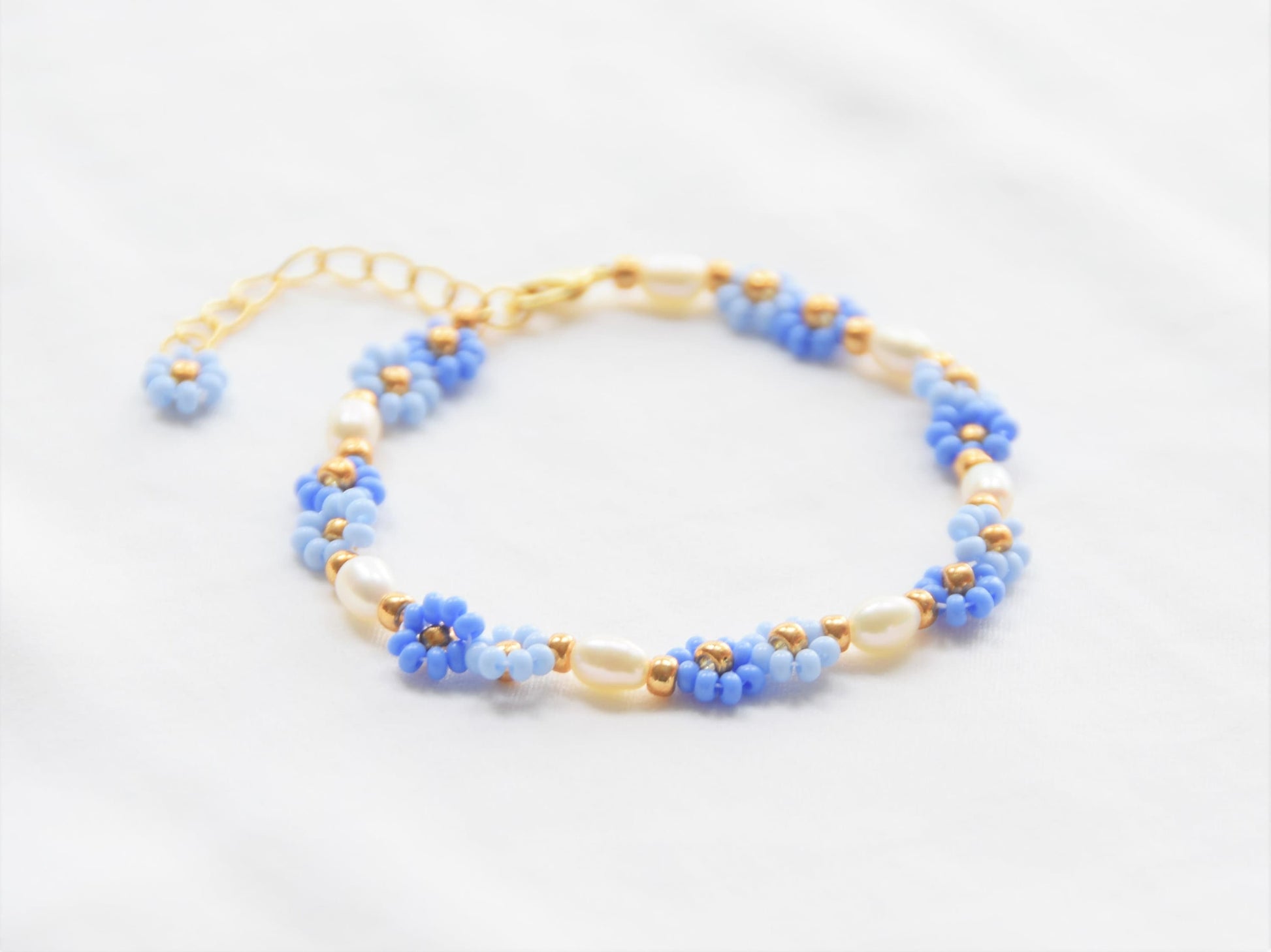 A bracelet made with seed beads and consists groups of two flowers in two shades of blue, alternating with oval shaped freshwater pearls. The clasp and extension chain are gold plated. A gold bead separates each group of two flowers and pearls.