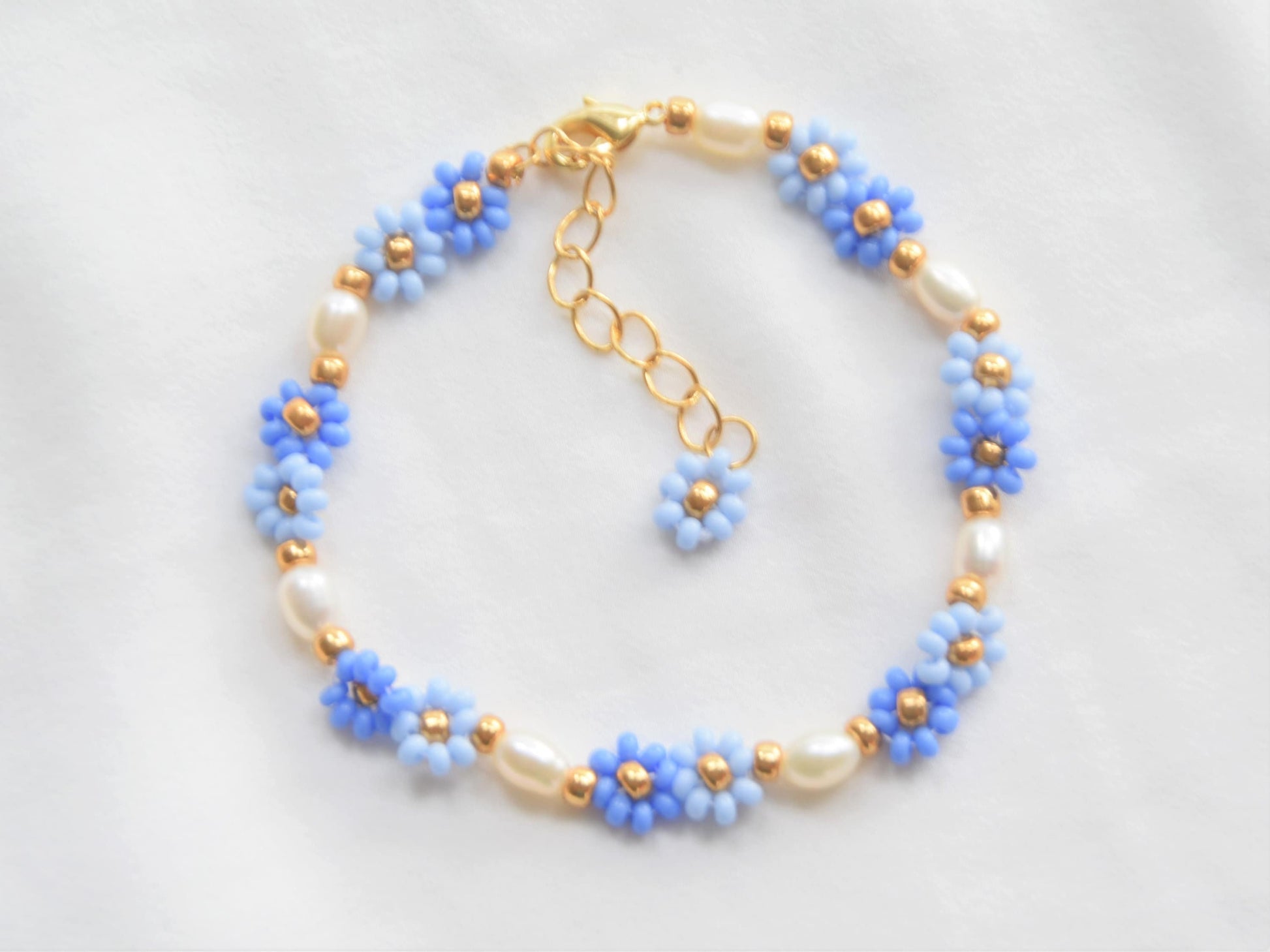 A bracelet made with seed beads and consists groups of two flowers in two shades of blue, alternating with oval shaped freshwater pearls. The clasp and extension chain are gold plated. A gold bead separates each group of two flowers and pearls.