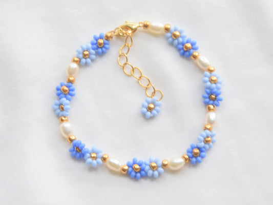 A bracelet made with seed beads and consists groups of two flowers in two shades of blue, alternating with oval shaped freshwater pearls. The clasp and extension chain are gold plated. A gold bead separates each group of two flowers and pearls.