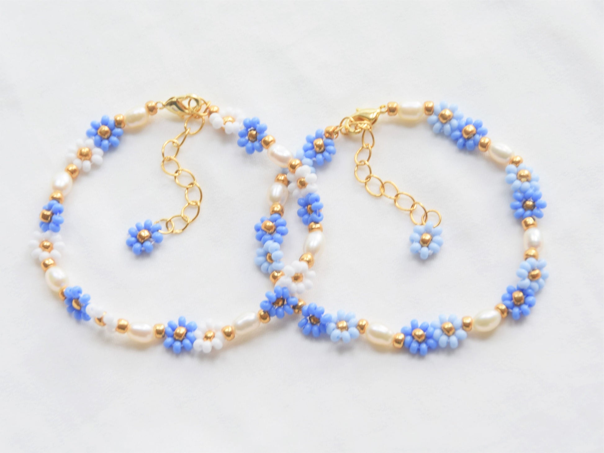 Two bracelets, side by side, slightly overlapping. One has blue and white flowers and the other one has two shades of blue flowers. The flowers are in groups of two, alternating with freshwater pearls. The clasps and extension chains are gold.