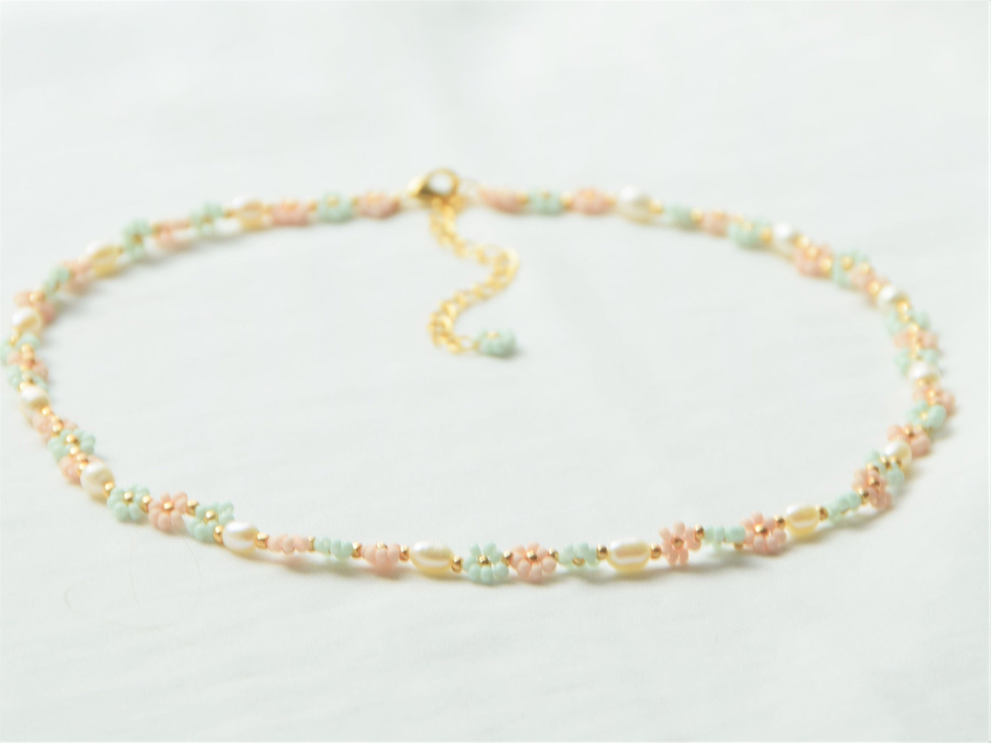 A necklace lying flat, making a circle, on a white background. The necklace has light green and light pink flowers, alternating each other in sets of three. Between each set of three flowers, is an oval shaped freshwater pearl.