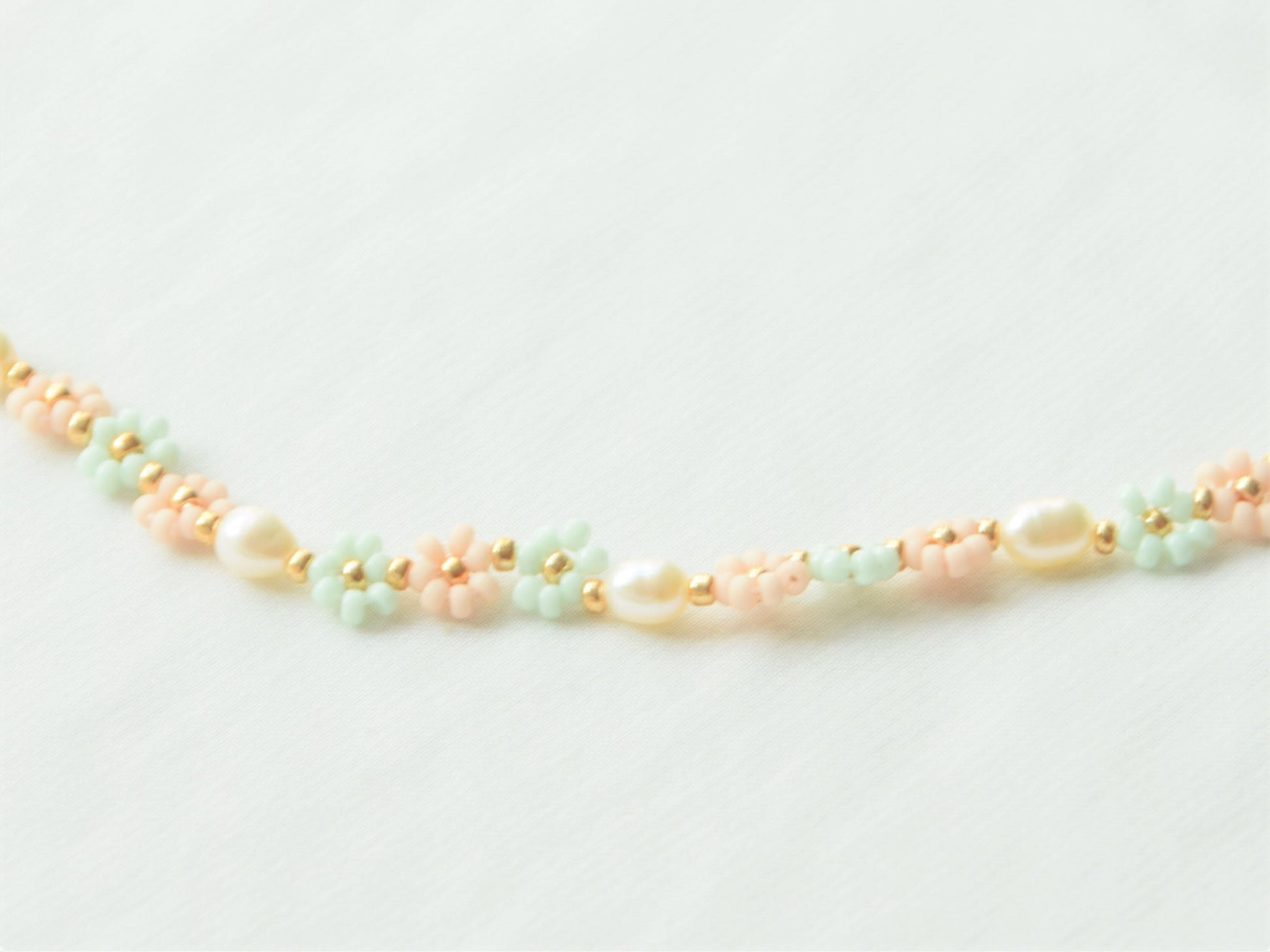 Close-up image of a necklace lying flat on a white background. The necklace has light green and light pink flowers, alternating each other in sets of three. Between each set of three flowers, is an oval shaped freshwater pearl.