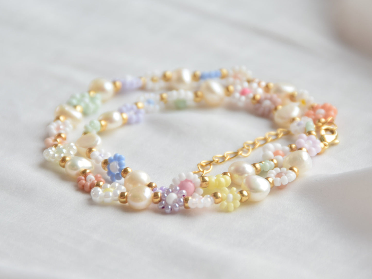 A necklace wrapped twice around itself, lying on a white surface. The necklace consists of small beaded flowers in many different soft colors. The flowers are in groups of three, two or one, alternating with culture pearls. The clasp is gold.