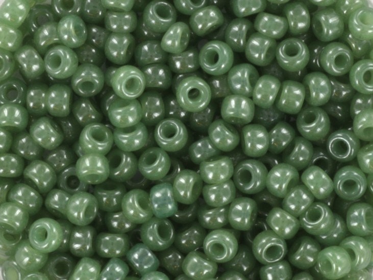 Translucent green seed beads, 3mm in size and round in shape. The image is a close-up of a bowl of full of seed beads so that the beads fill the entire image