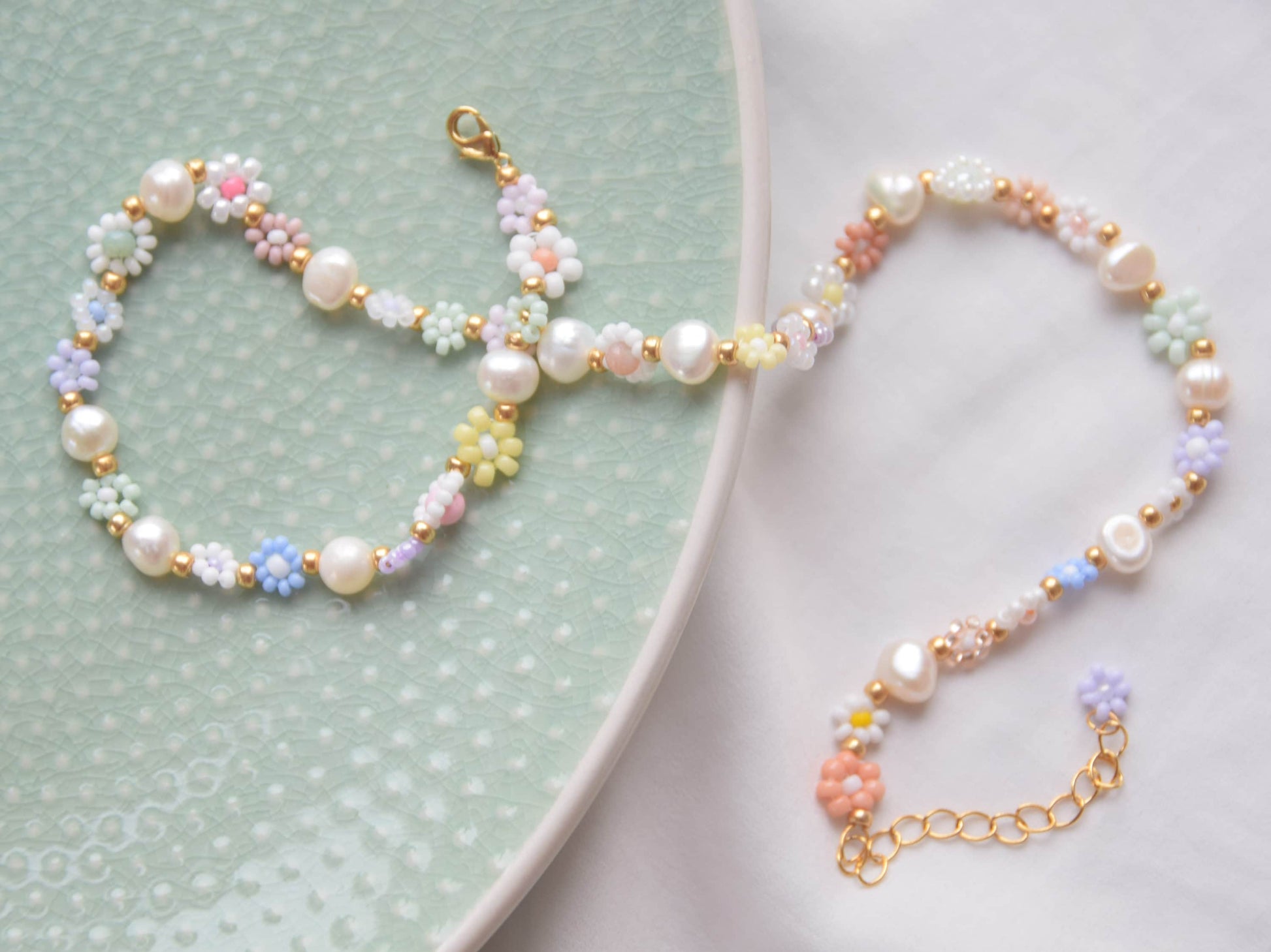 A necklace lying, half on a light green plate, half on a white surface. The necklace consists of small beaded flowers in many different soft colors. The flowers are in groups of three, two or one, alternating with culture pearls. The clasp is gold.