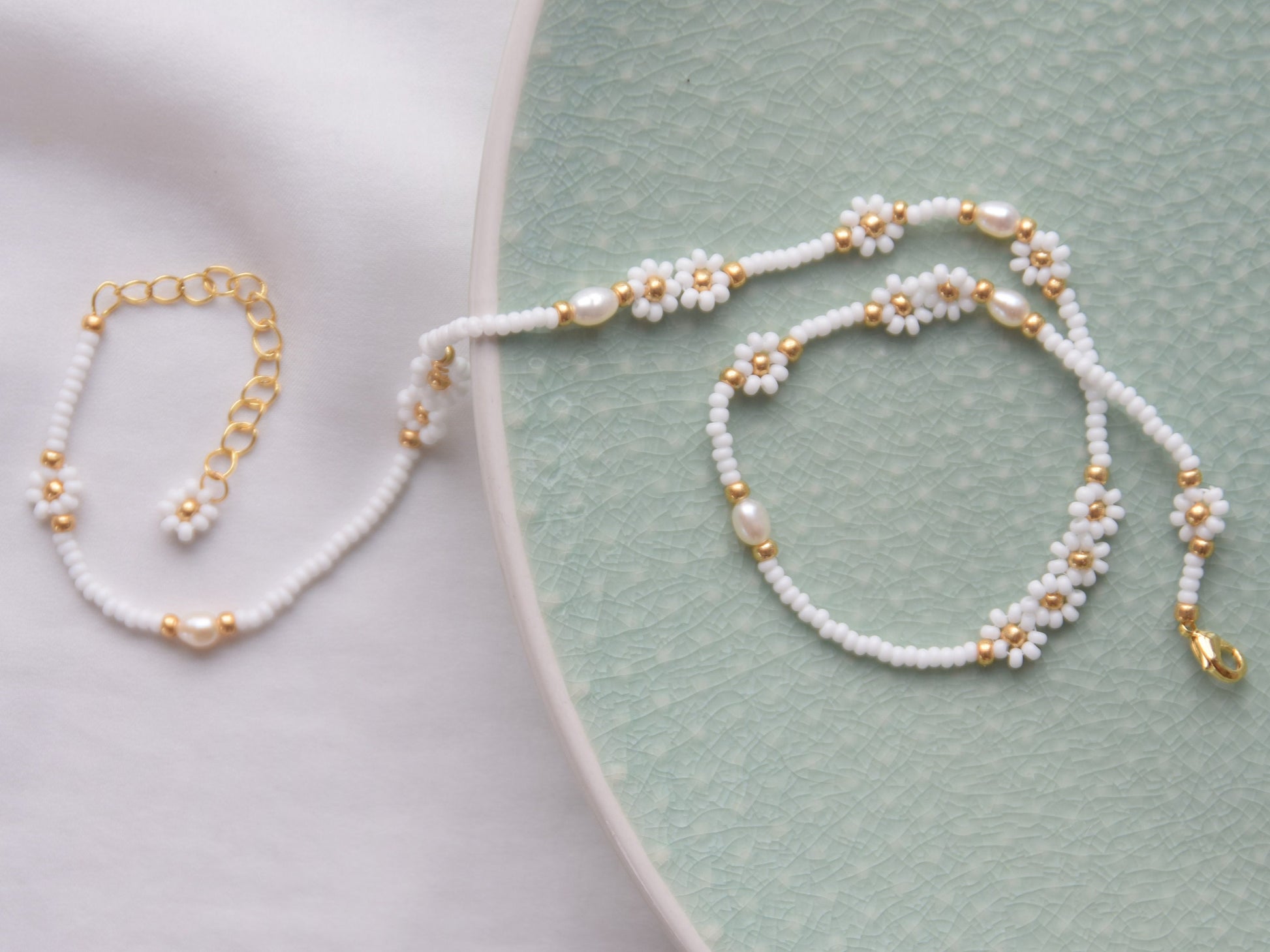 A single stranded white beaded necklace. The beads are arranged in a single line and some are arranged in little flowers with white petals and a gold center. Between the flowers and single stranded beads, occasionally is a freshwater pearl.