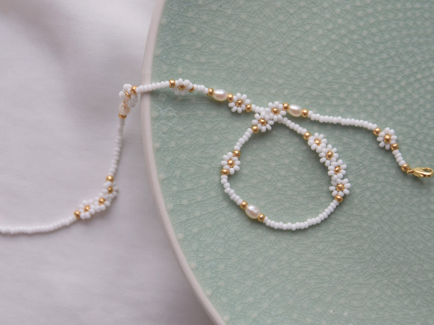 A single stranded white beaded necklace. The beads are arranged in a single line and some are arranged in little flowers with white petals and a gold center. Between the flowers and single stranded beads, occasionally is a freshwater pearl.