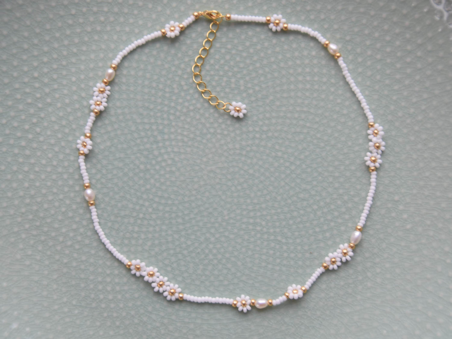 A single stranded white beaded necklace. The beads are arranged in a single line and some are arranged in little flowers with white petals and a gold center. Between the flowers and single stranded beads, occasionally is a freshwater pearl.