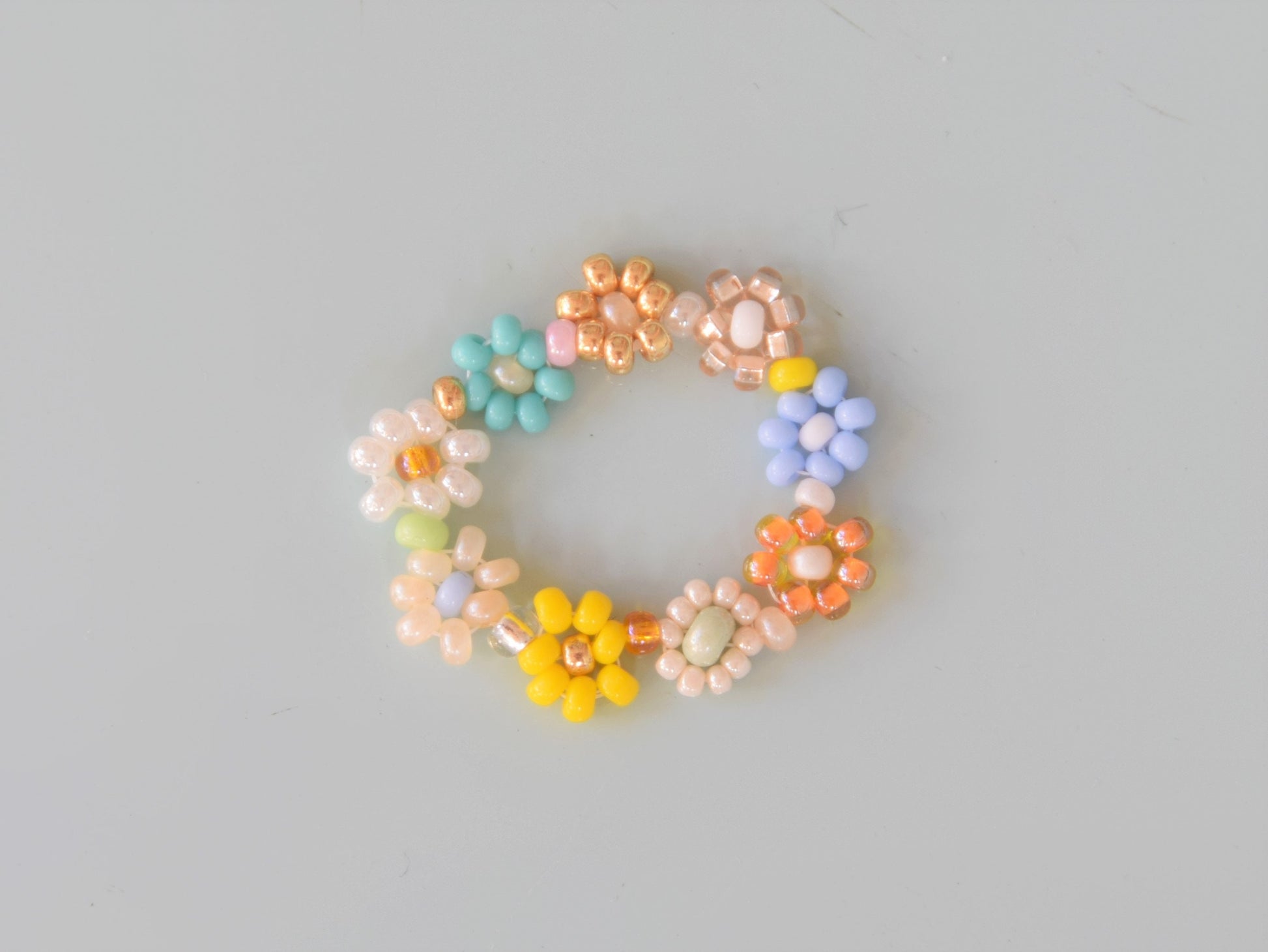 A colorful ring laying flat on a light blue surface. The ring consists of small round glass beads of several different colors, arranged into flowers.