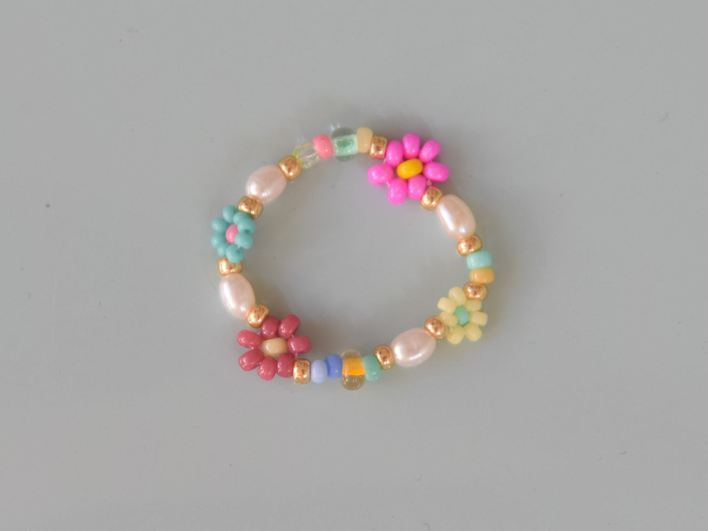 Colorful ring laying on a light blue surface. The ring consists of small round glass beads and oval shaped freshwater pearls. Some of the glass beads are arranged into flowers. There are gold colored glass beads on each end of the pearls and flowers