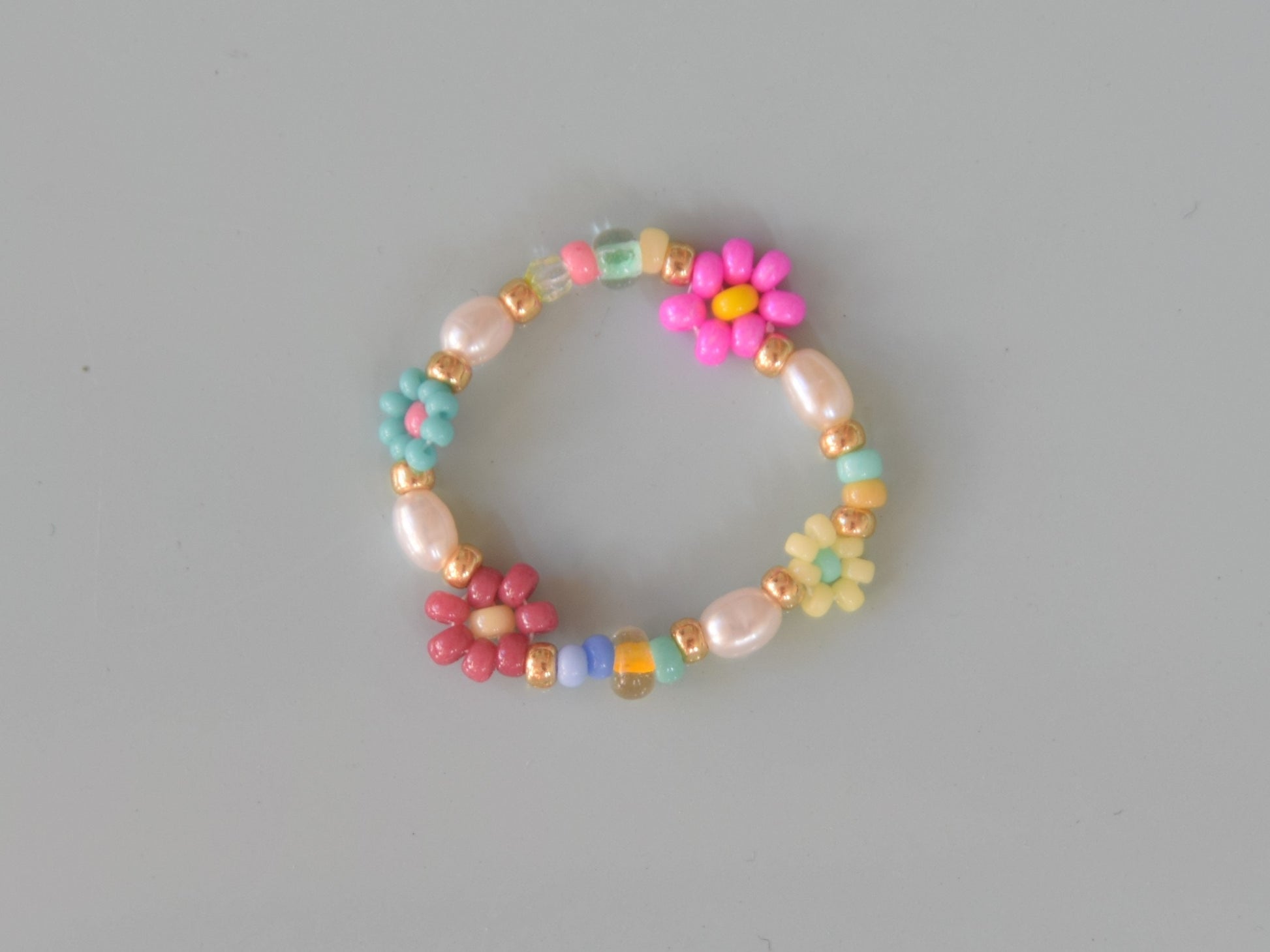 Colorful ring laying on a light blue surface. The ring consists of small round glass beads and oval shaped freshwater pearls. Some of the glass beads are arranged into flowers. There are gold colored glass beads on each end of the pearls and flowers