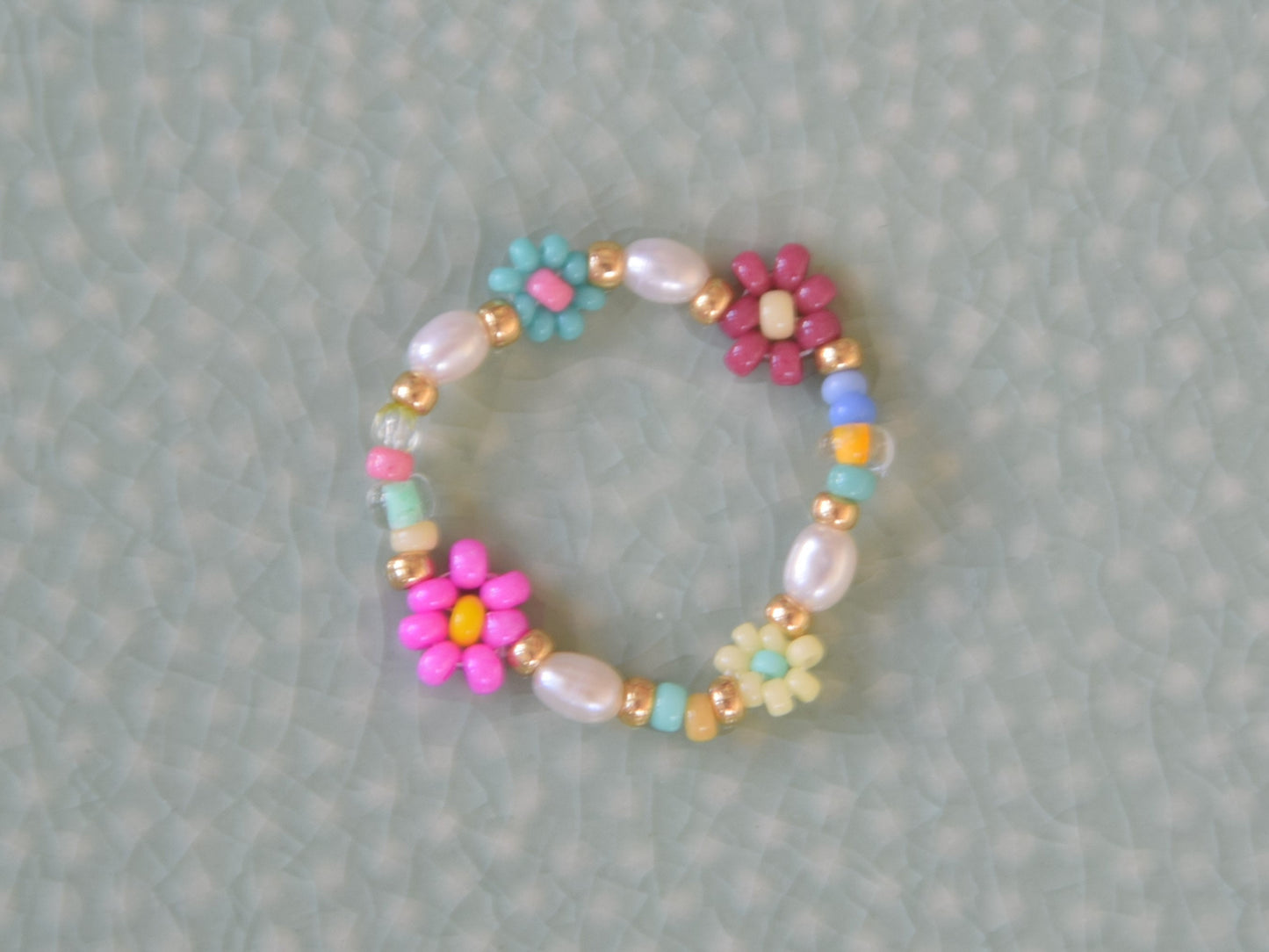 Colorful ring laying on a light green surface. The ring consists of small round glass beads and oval shaped freshwater pearls. Some of the glass beads are arranged into flowers. There are gold colored glass beads on each end of the pearls and flowers