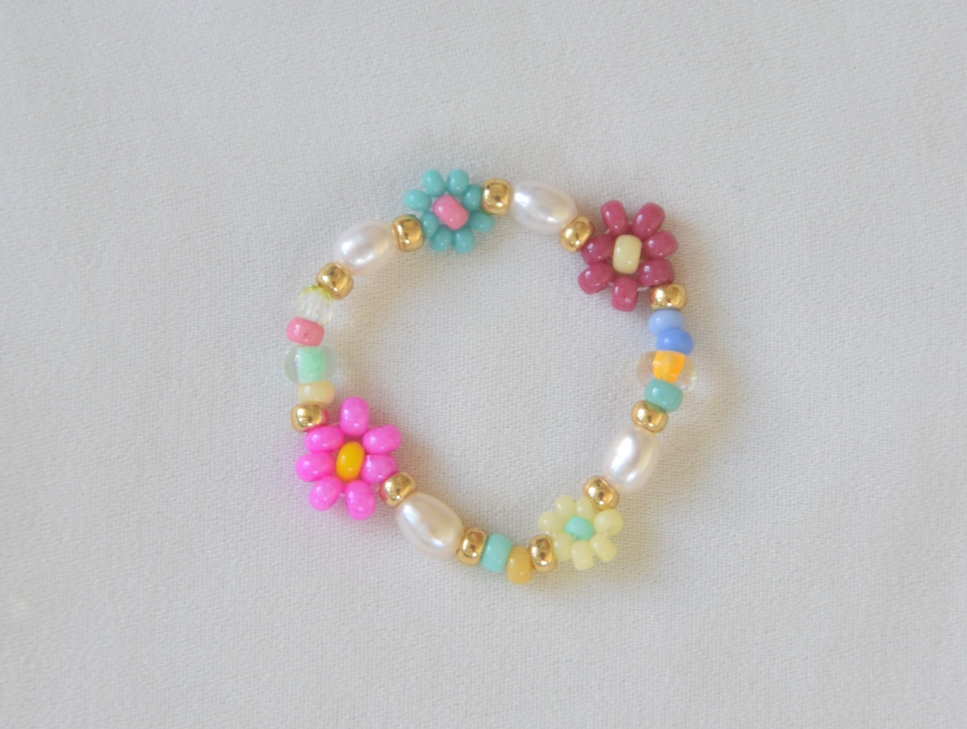 Colorful ring laying on a white surface. The ring consists of small round glass beads and oval shaped freshwater pearls. Some of the glass beads are arranged into flowers. There are gold colored glass beads on each end of the pearls and the flowers