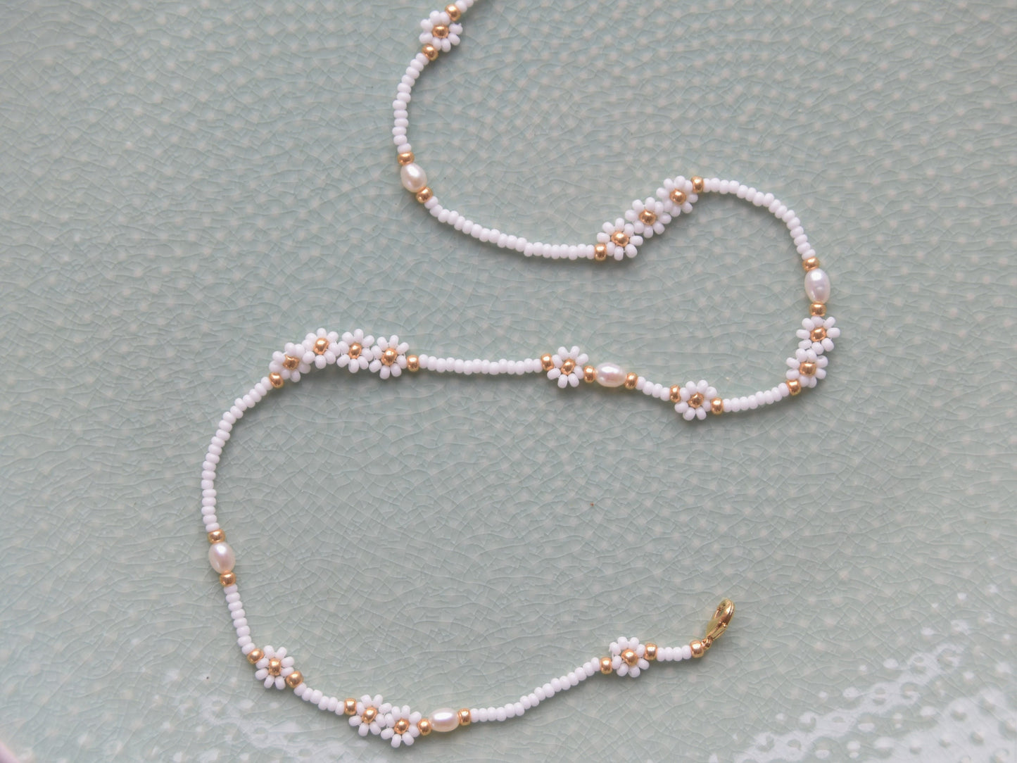 A single stranded white beaded necklace. The beads are arranged in a single line and some are arranged in little flowers with white petals and a gold center. Between the flowers and single stranded beads, occasionally is a freshwater pearl.