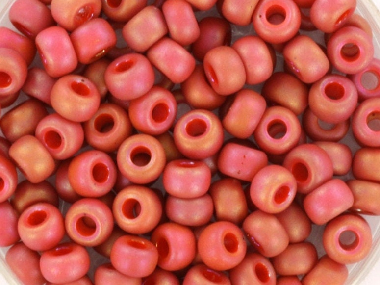 Red seed beads with rainbow luster, 4mm in size and round in shape. The image is a close-up of a bowl of full of seed beads so that the beads fill the entire image