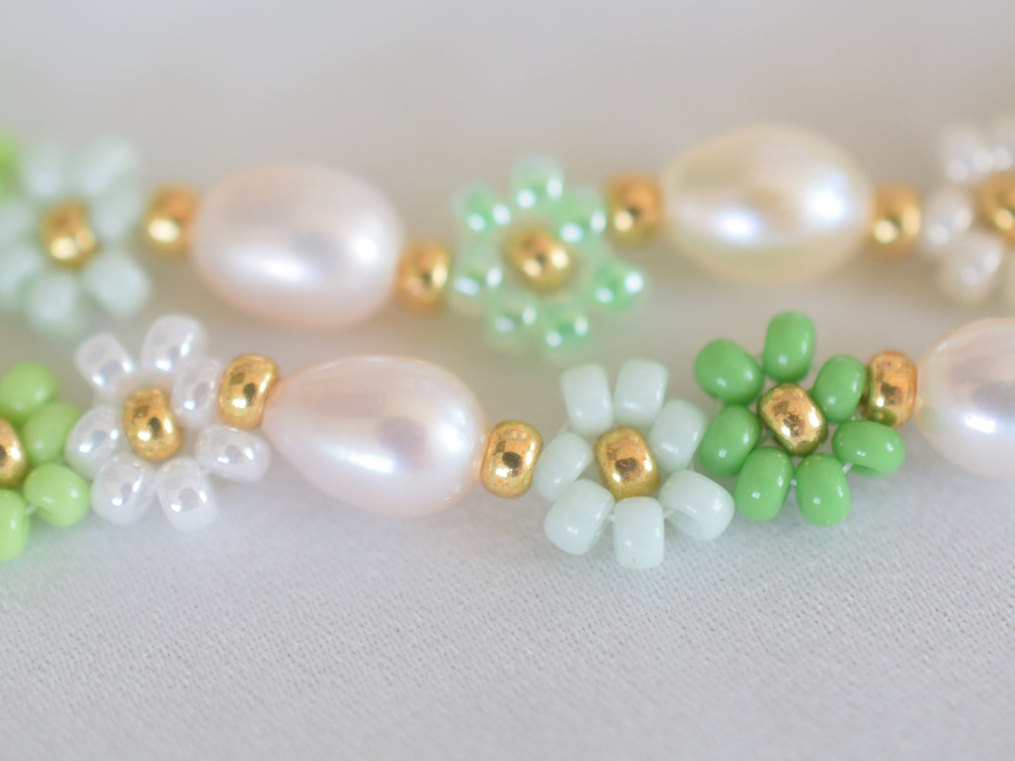 Beaded necklace with seed beads and oval freshwater pearls. The seed beads are in various shades of green and white, arranged in little flowers with a gold bead as the center of each flower. The necklace has a gold colored lobster clasp.