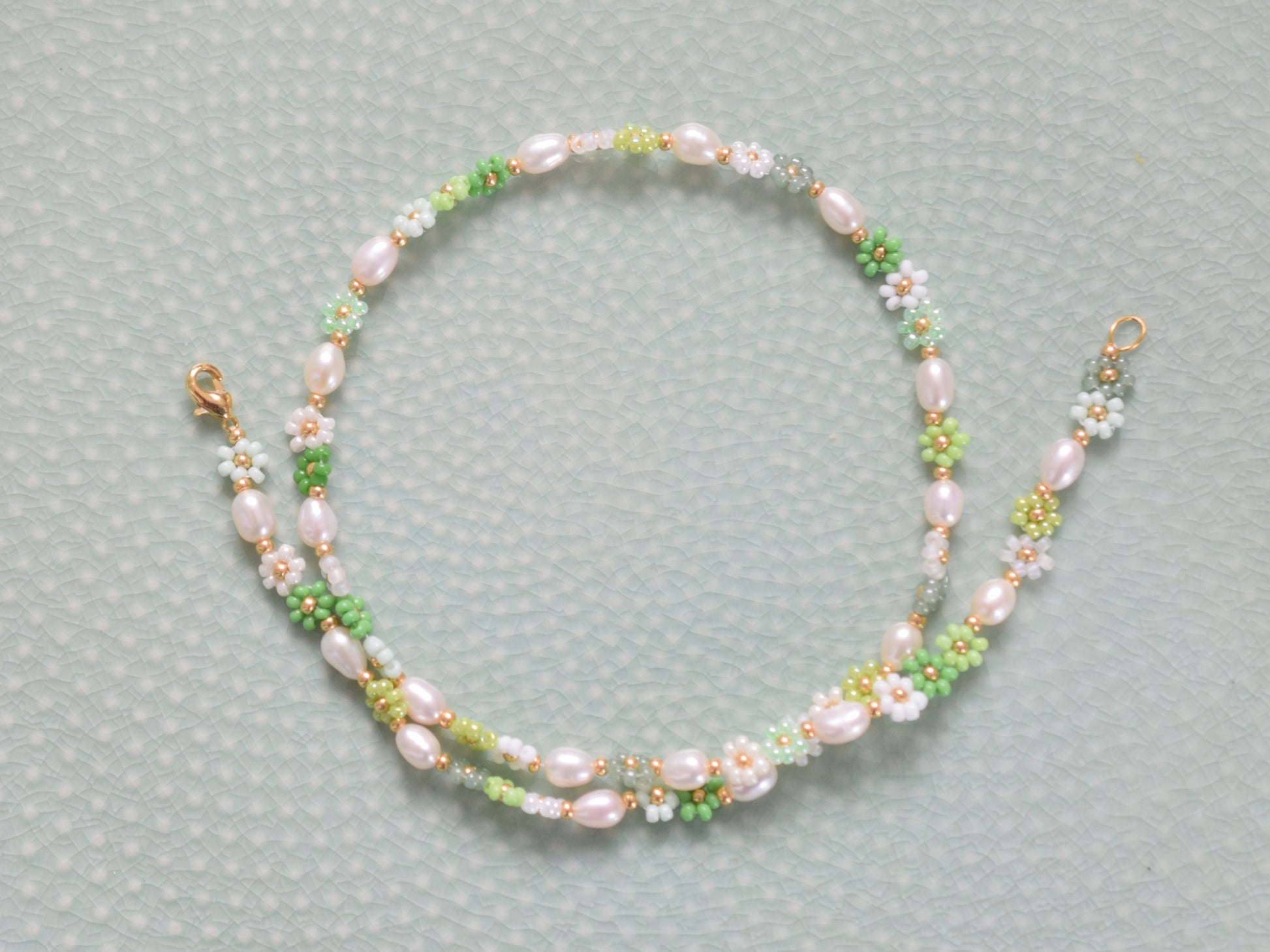 Beaded necklace with seed beads and oval freshwater pearls. The seed beads are in various shades of green and white, arranged in little flowers with a gold bead as the center of each flower. The necklace has a gold colored lobster clasp.