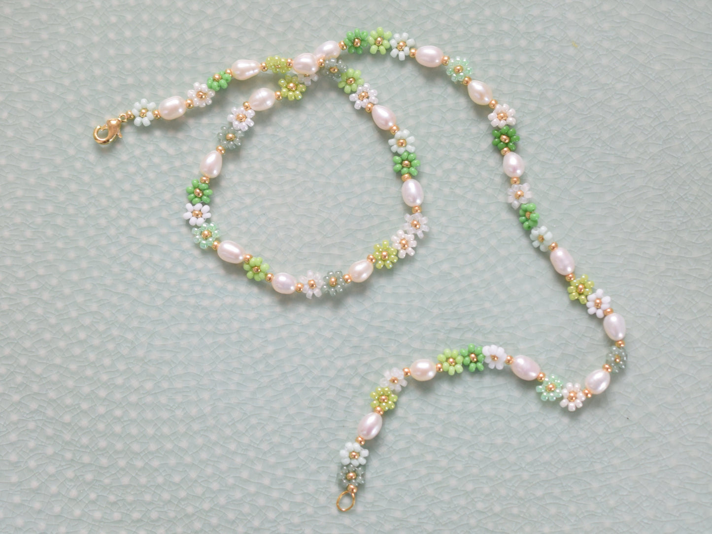 Beaded necklace with seed beads and oval freshwater pearls. The seed beads are in various shades of green and white, arranged in little flowers with a gold bead as the center of each flower. The necklace has a gold colored lobster clasp.