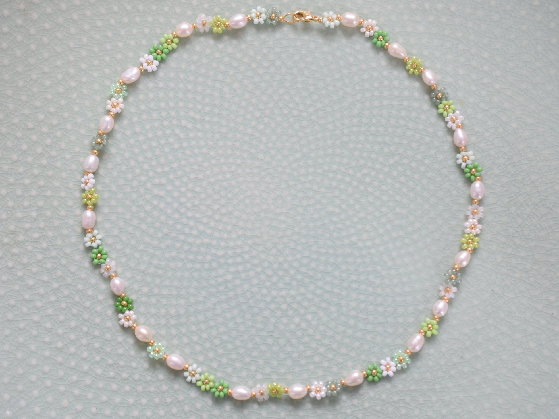 Beaded necklace with seed beads and oval freshwater pearls. The seed beads are in various shades of green and white, arranged in little flowers with a gold bead as the center of each flower. The necklace has a gold colored lobster clasp.