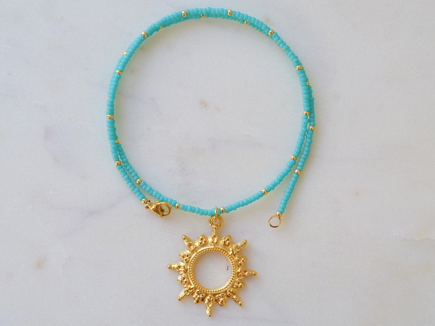 A turquoise beaded necklace with small gold spacers, featuring a gold sunburst pendant in the center. The necklace is laid out in a circular shape on a light, textured background. A clasp is visible on one end.