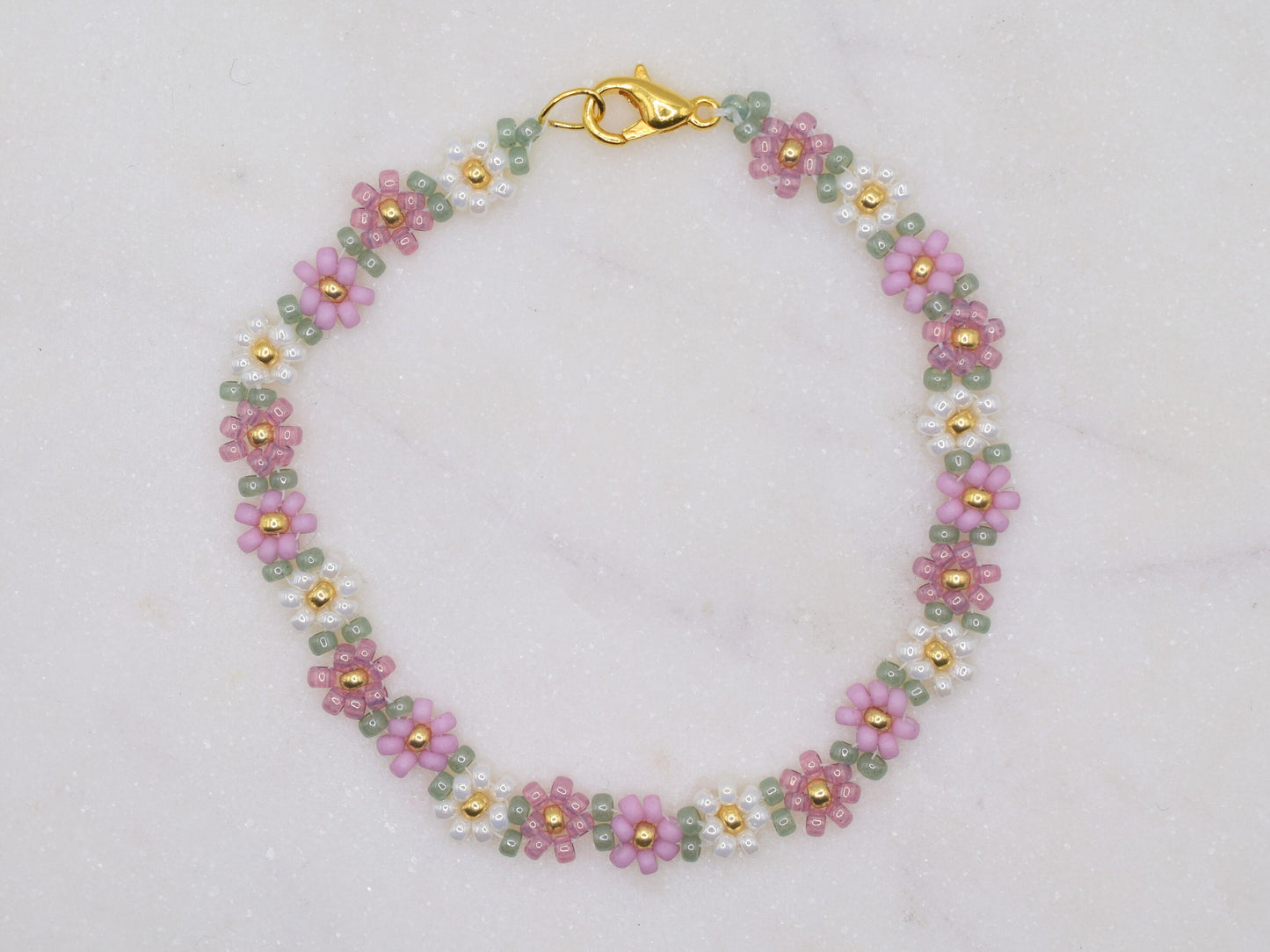 A beaded bracelet featuring small floral patterns in pink, green, and white beads with gold accents. The bracelet is arranged in a circular shape on a light, textured background. A gold clasp is visible at one end.