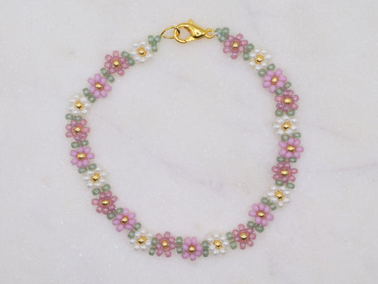 A beaded bracelet featuring small floral patterns in pink, green, and white beads with gold accents. The bracelet is arranged in a circular shape on a light, textured background. A gold clasp is visible at one end.