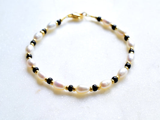 A delicate bracelet featuring oval freshwater pearls interspersed with small black beads and gold spacers. The pearls are white with a subtle sheen, and the bracelet is secured with a gold clasp. The design is elegant and timeless.