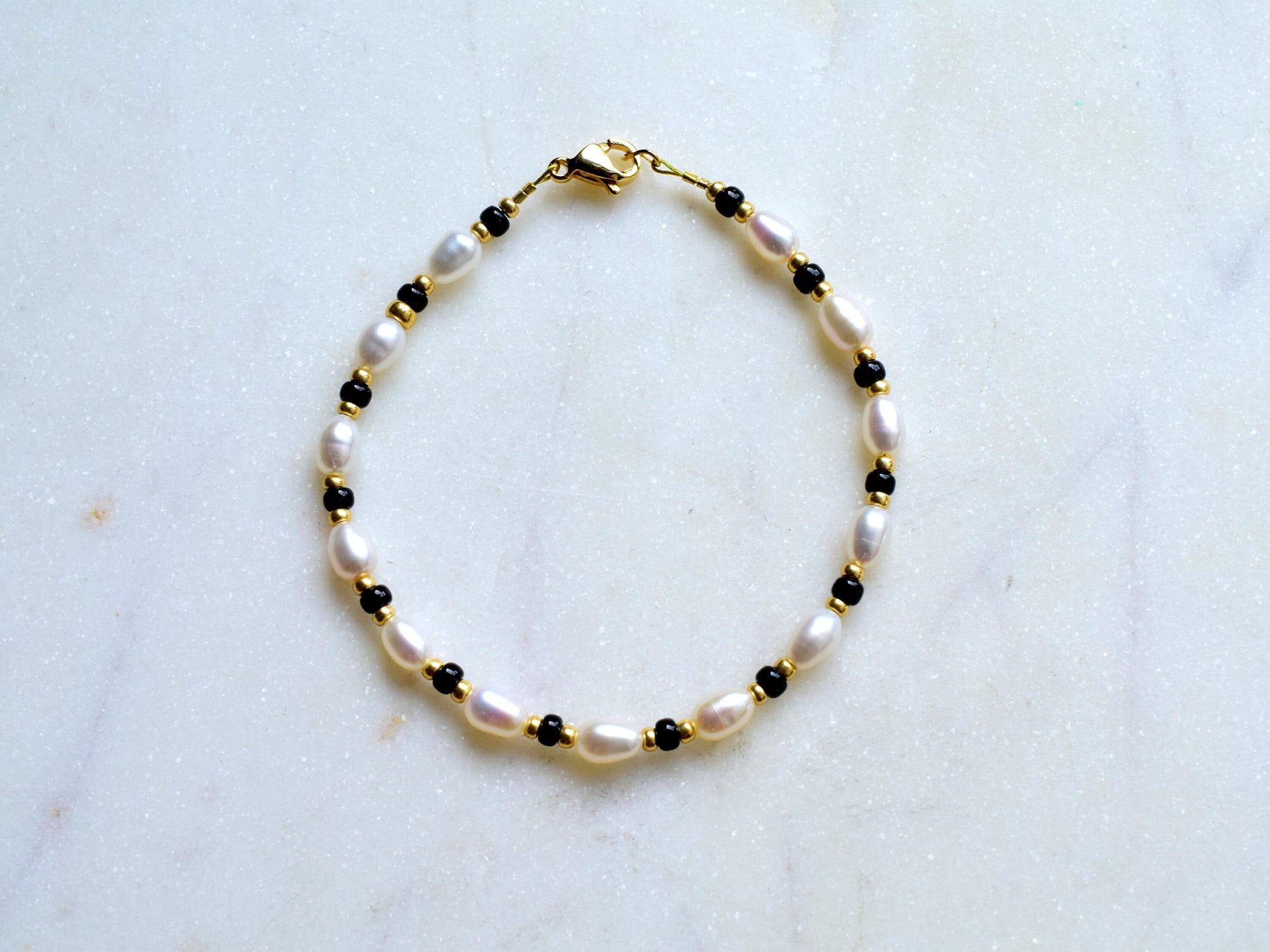 A delicate bracelet featuring oval freshwater pearls interspersed with small black beads and gold spacers. The pearls are white with a subtle sheen, and the bracelet is secured with a gold clasp. The design is elegant and timeless.